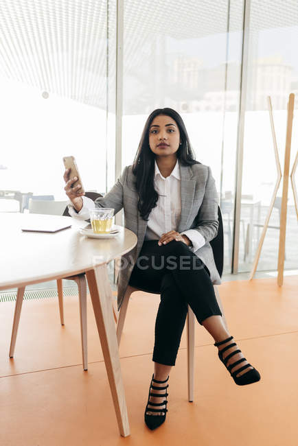 Elegante Geschäftsfrau mit Smartphone, die mit einer Tasse Tee am Tisch sitzt und in die Kamera blickt — Stockfoto