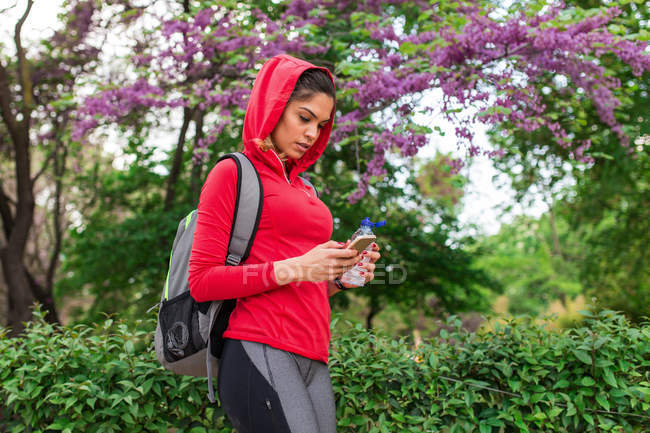 Portrait à angle bas de fille sportive naviguant smartphone sur arbre en fleurs au parc — Photo de stock