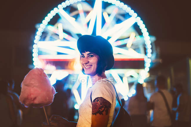Mulher elegante posando com nuvem de açúcar e olhando para a câmera no fundo da roda de balsa iluminada — Fotografia de Stock