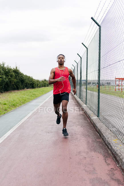 Vista frontal do homem usando fones de ouvido correndo na pista ao longo da cerca — Fotografia de Stock