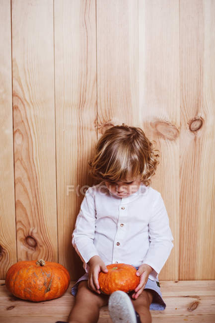 Kleines Kind sitzt auf Holzuntergrund und blickt nachdenklich auf Kürbisse — Stockfoto