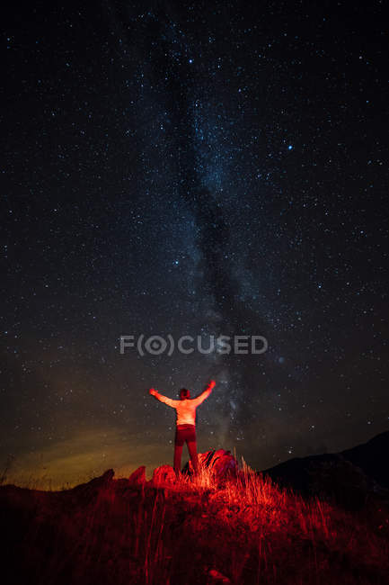 Rückansicht eines rotbeleuchteten Mannes, der mit erhobenen Armen unter Milchstraße im Himmel posiert — Stockfoto