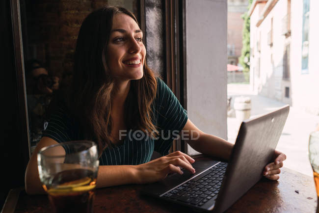 Retrato de mujer alegre sentada con portátil en la cafetería y mirando a un lado - foto de stock