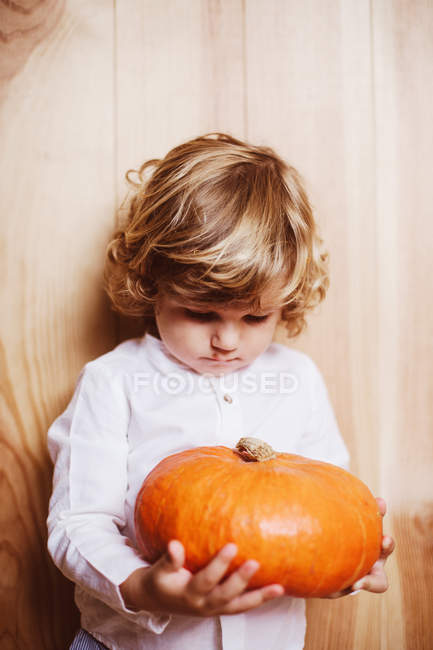 Menino adorável sentado à beira da parede de madeira e olhando penosamente na abóbora nas mãos — Fotografia de Stock