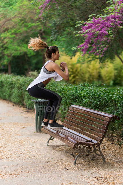 Side view of sporty girl jumping on bench in park — Stock Photo