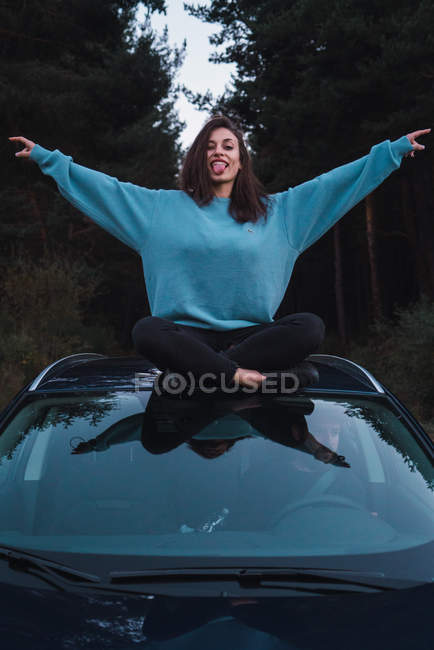 Brünettes Mädchen in blauem Sweatshirt sitzt mit erhobenen Armen auf dem Auto und zeigt Zunge in die Kamera — Stockfoto