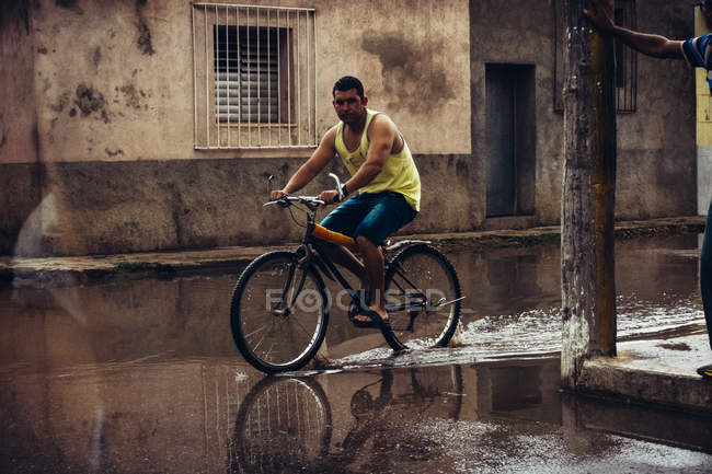 CUBA - AUGUST 27, 2016: Side view of man riding bicycle through paddle on background of poor city district. — Stock Photo