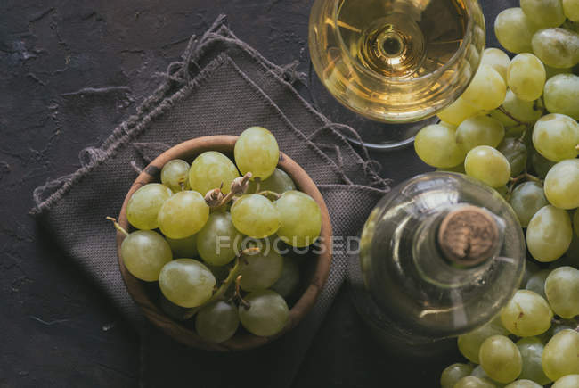 Vista directa del racimo de uvas verdes en un tazón al lado de un vaso de vino y una botella en la mesa - foto de stock