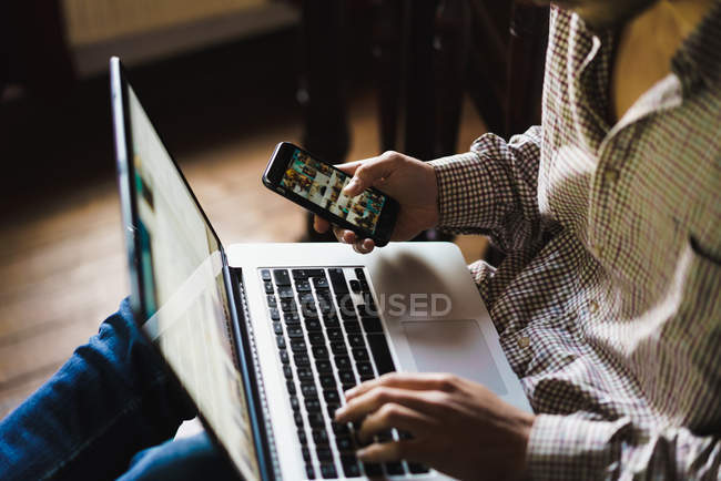 Кроп человек сидит на деревянной лестнице с ноутбуком на коленях и с помощью смартфона — стоковое фото