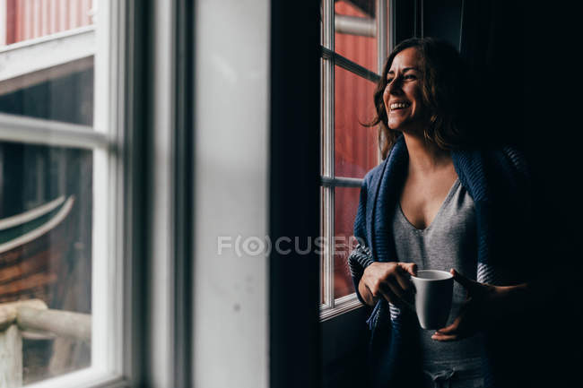 Mujer sonriente con taza mirando en la ventana - foto de stock