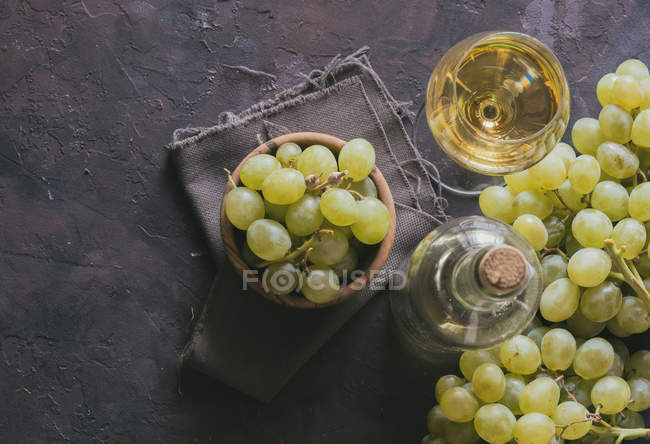 Vista superior de cacho de uvas verdes na tigela ao lado de copo de vinho e garrafa na mesa — Fotografia de Stock