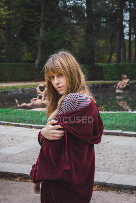 Chica morena en chaqueta roja posando junto a la fuente barroca y mirando por encima del hombro a la cámara - foto de stock