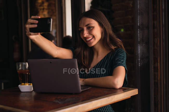 Mujer sonriente sentada en la cafetería con portátil en la mesa y tomando selfie - foto de stock