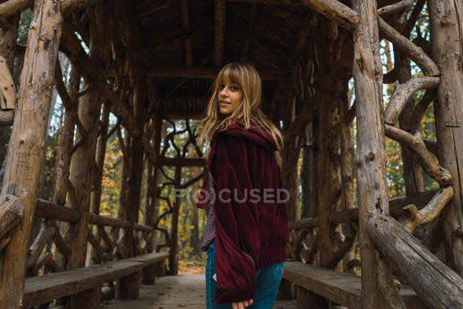 Brunette girl in red hoodie jacket walking in wooden passage and looking over shoulder at camera — Stock Photo