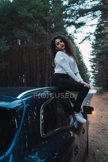 Brunette femme assise sur le dessus de la voiture garée sur le côté de la route forestière . — Photo de stock