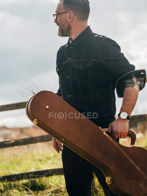 Вид сбоку на человека в джинсовой рубашке, позирующего с гитарным чехлом на поле — стоковое фото