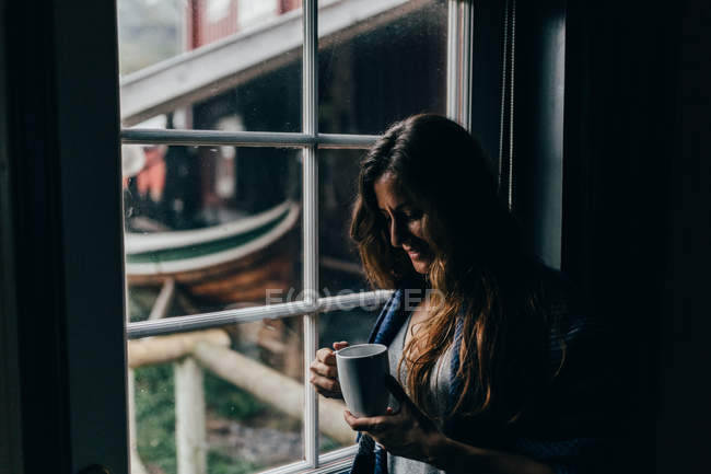 Mulher atraente de pé perto da janela e olhando em caneca de café — Fotografia de Stock