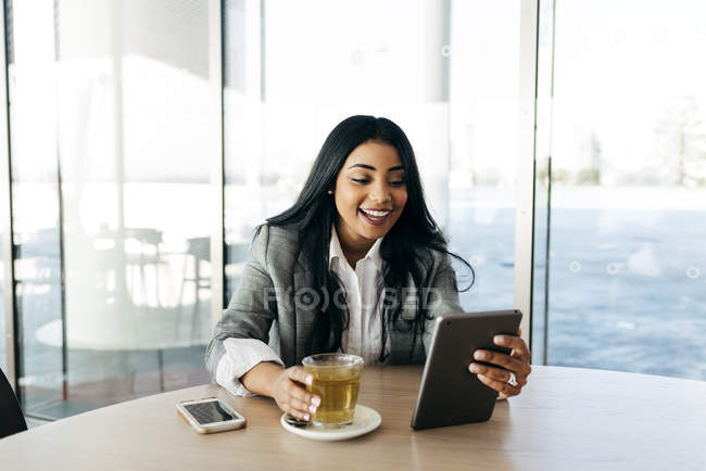 Fröhliche stilvolle Geschäftsfrau trinkt Tee und blickt auf Tablette in der Hand. — Stockfoto