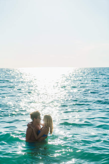 Vista lateral do casal abraçando posando na água do oceano com linha de luz do sol — Fotografia de Stock