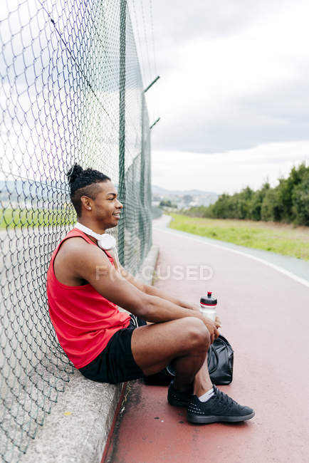 Vista lateral del atleta sentado cerca de la valla y descansando después del entrenamiento - foto de stock