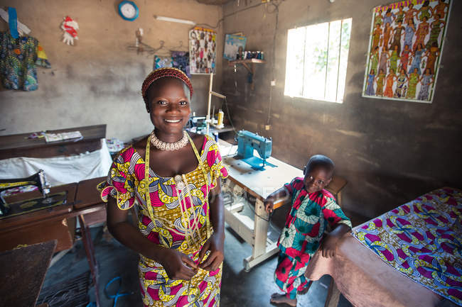 BENIN, ÁFRICA - 31 de agosto de 2017: Mujer alegre con su hijo sonriendo de pie en el taller y mirando a la cámara . - foto de stock