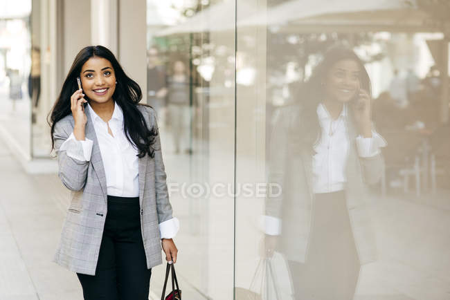 Усміхнена бізнес-леді ходить по вікнах магазинів і розмовляє по телефону — стокове фото