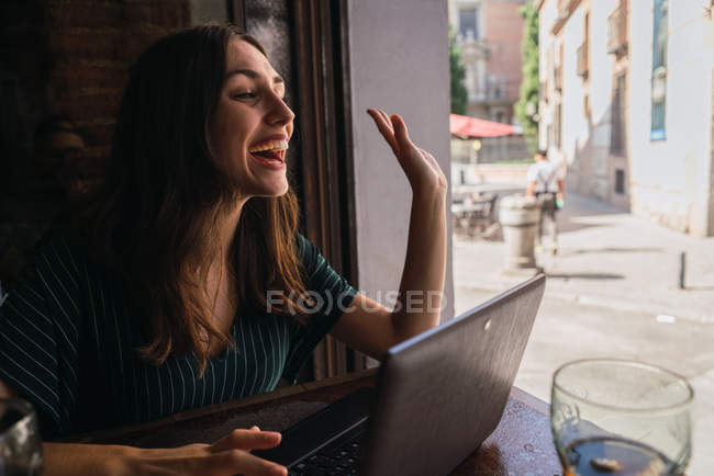 Smiling woman sitting with laptop in cafe looking aside and greeting with gesture — Stock Photo