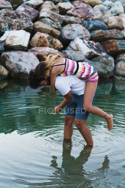 Side view of man carrying girlfriend on back and walking in shallow coastline water. — Stock Photo
