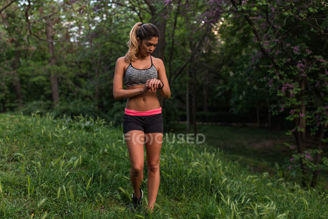 Deportiva chica posando en el césped del parque y mirando reloj en el brazo - foto de stock