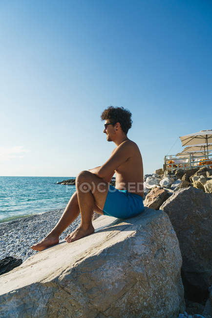 Side view of man in sunglasses and swimming shorts sitting on boulder and looking at ocean. — Stock Photo