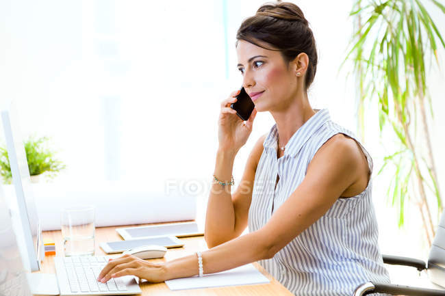 Retrato de mulher de negócios usando laptop e falando no smartphone no escritório moderno . — Fotografia de Stock