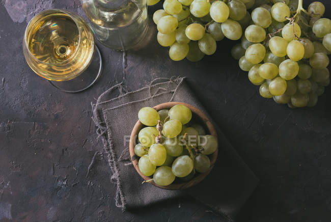 Вид сверху на пучок зеленого винограда со шкурками в миске рядом с бокалом белого вина на столе — стоковое фото