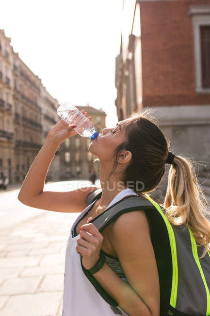 Вид сбоку на девушку в спортивной одежде, стоящую с рюкзаком на улице и питьевой водой . — стоковое фото