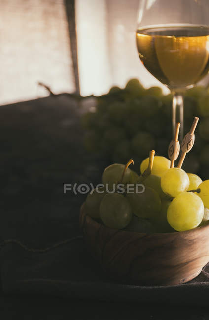 Vista de perto de cacho de uvas verdes com espetos em tigela no fundo de copo de vinho branco — Fotografia de Stock