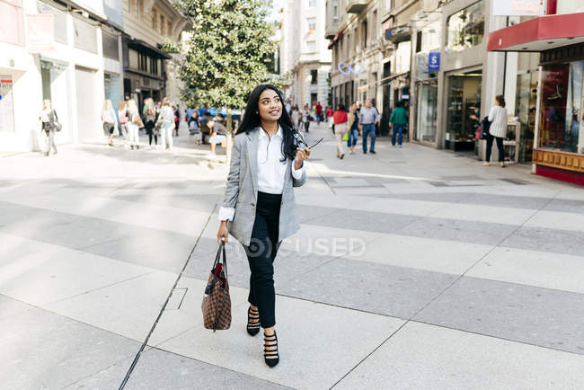 Elegante Geschäftsfrau, die mit Handy und Handtasche in der Hand auf der Straße spaziert und nach oben schaut — Stockfoto