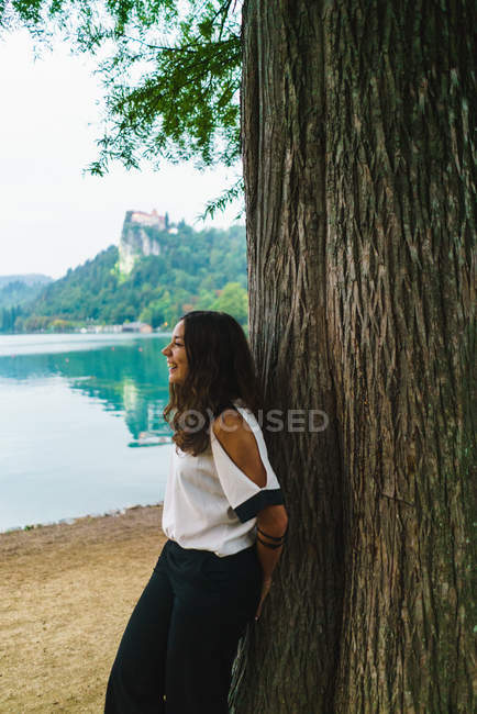 Seitenansicht eines brünetten Mädchens, das sich am Ufer des Sees an einen Baumstamm lehnt — Stockfoto
