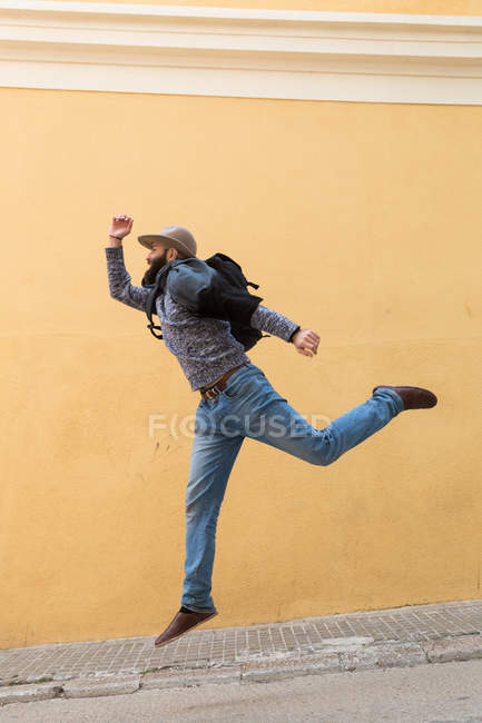 Turista barbudo con mochila saltando en la pared amarilla de la calle . - foto de stock