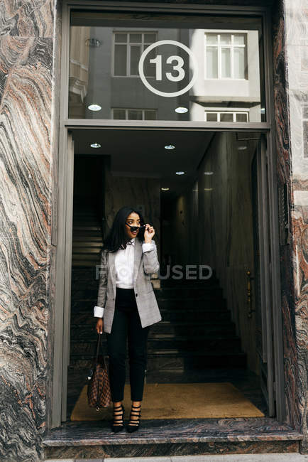 Portrait of elegant businesswoman walking out of building and putting on sunglasses. — Stock Photo