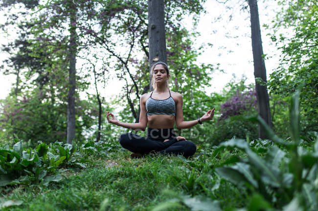 Low angle view of sporty woman sitting on ground and meditating in park — Stock Photo