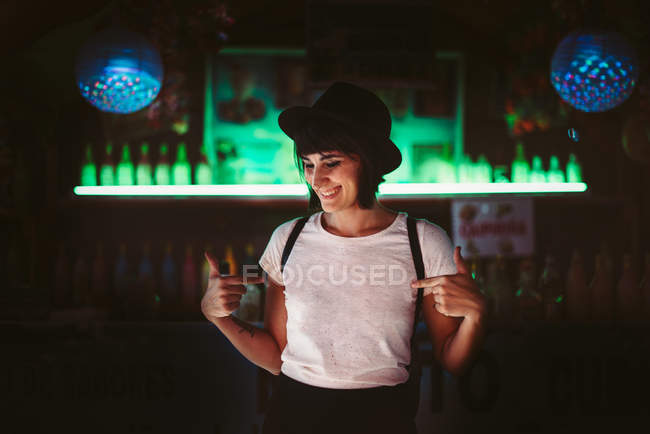 Mulher elegante jovem apontando para si mesma em bar escuro com prateleiras iluminadas . — Fotografia de Stock