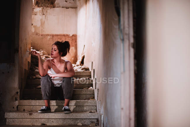 Mujer joven sentada en las escaleras de la casa envejecida y fumando cigarrillo - foto de stock