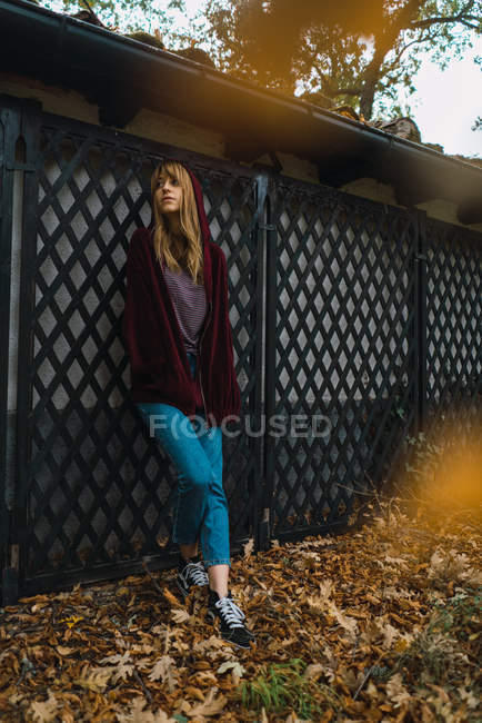 Brünettes Mädchen mit Kapuze lehnt romantisch an Zaun im Park und schaut weg. — Stockfoto