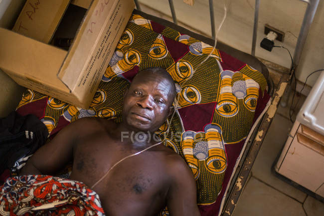 BENIN, ÁFRICA - 31 de agosto de 2017: Vista superior del hombre acostado en la cama en el hospital africano y mirando a la cámara - foto de stock