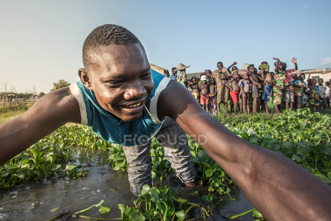 BENIN, ÁFRICA - 31 de agosto de 2017: Retrato de un hombre étnico sonriente que se inclina ante la cámara con las manos extendidas sobre el fondo de un grupo de personas en la orilla del estanque - foto de stock