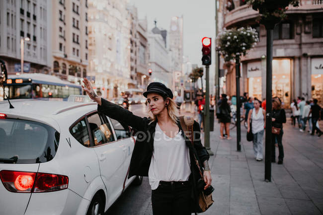 Young woman standing on sidewalk with hand up gesturing to taxi. — Stock Photo