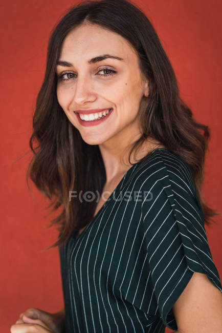 Porträt einer fröhlichen brünetten Frau im gestreiften Kleid, die vor rotem Hintergrund in die Kamera lächelt — Stockfoto