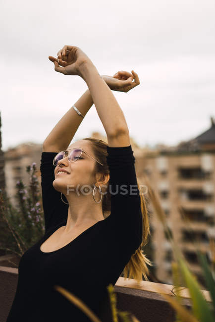 Ritratto di giovane bruna in occhiali in posa con le braccia alzate in terrazza — Foto stock