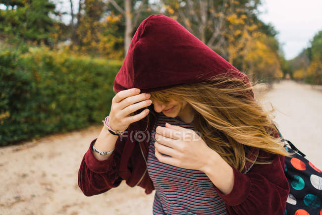 Brunette girl wearing hood and covering face from strong wind while walking in park. — Stock Photo