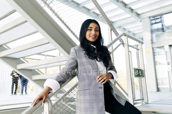 Mujer de negocios con chaqueta apoyada en pasamanos en el pasillo de las escaleras y mirando a la cámara - foto de stock