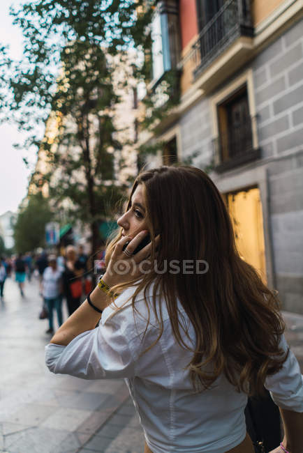 Rear view of young brunette speaking on smartphone and looking away at street scene — Stock Photo